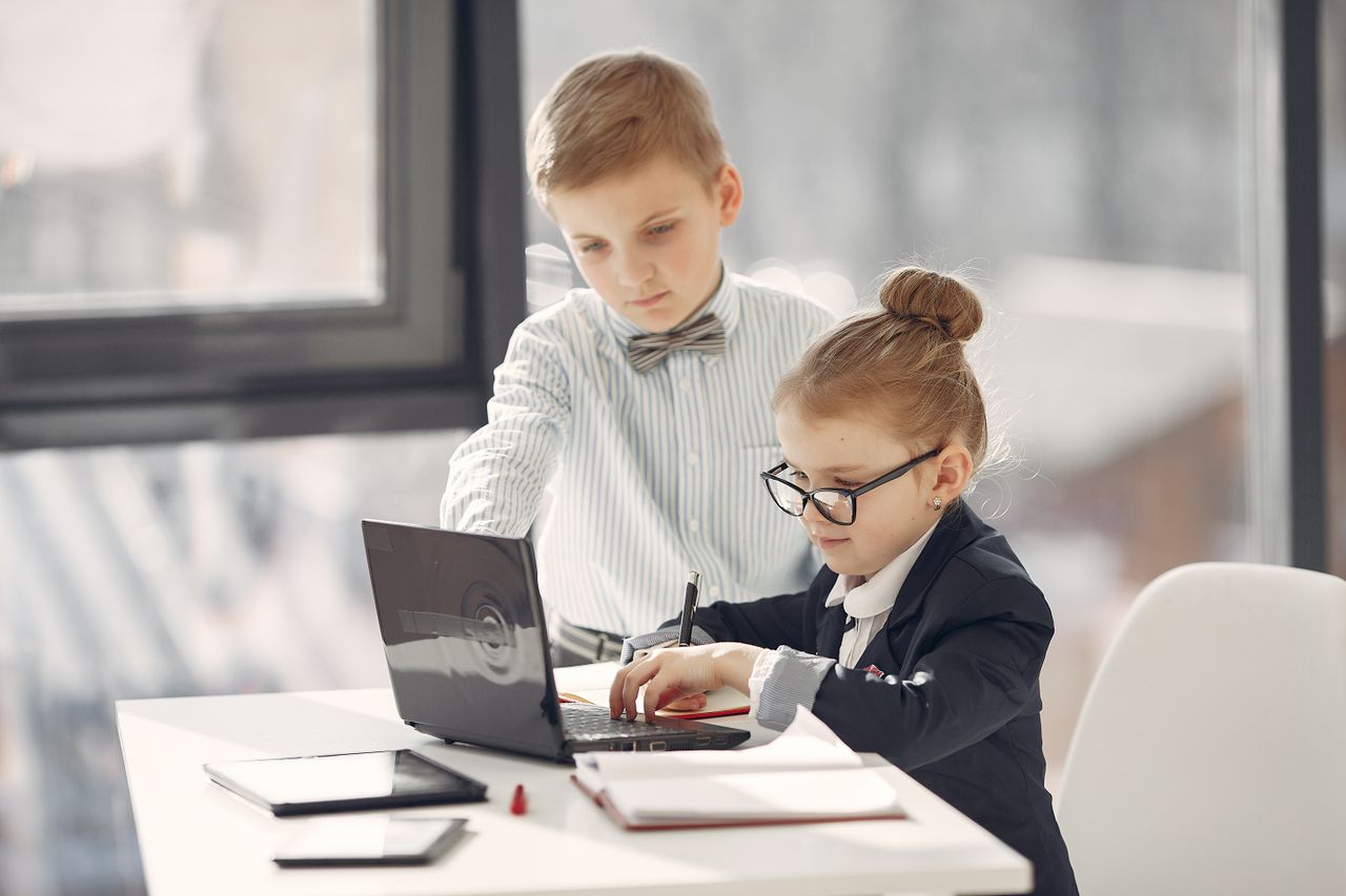 Focused boy wearing striped shirt and bow tie browsing netbook with smart little girl wearing formal suit and glasses and taking notes in notebook while discussing project during office work near table with different gadgets