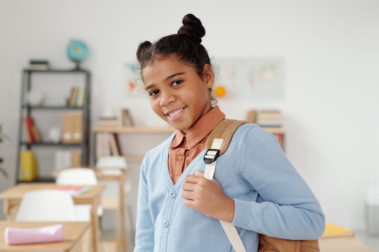 Smiling schoolgirl wearing a blue sweater stands in a classroom, ready for school.