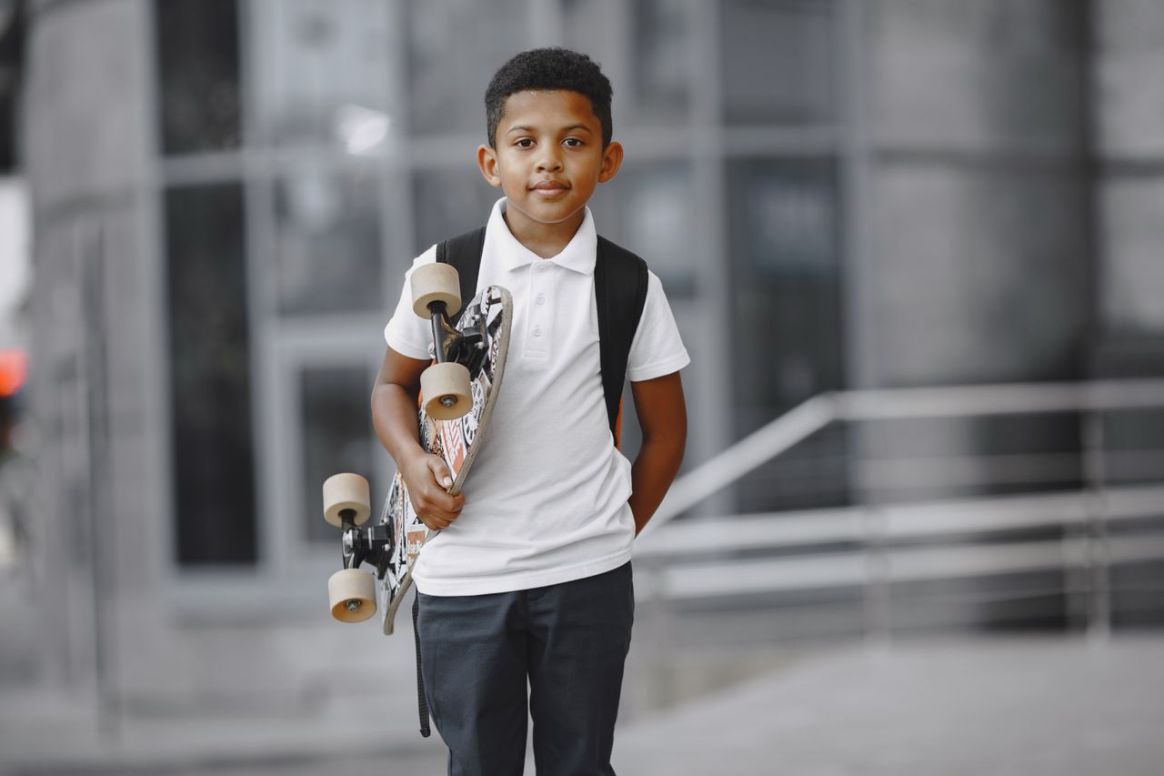 A young boy holding a skateboard and wearing a backpack, standing outside.