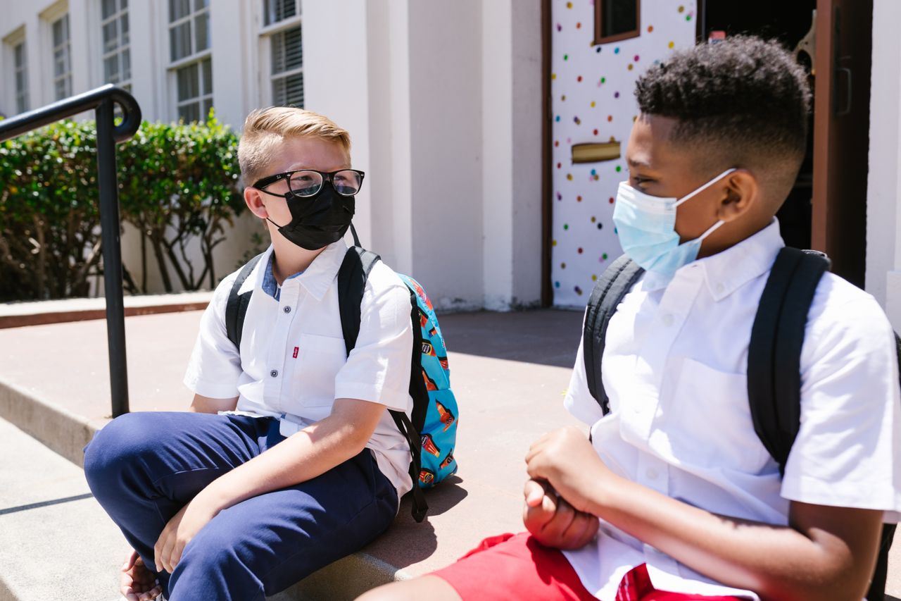 Two kids in masks sitting outside school, enjoying social interaction.