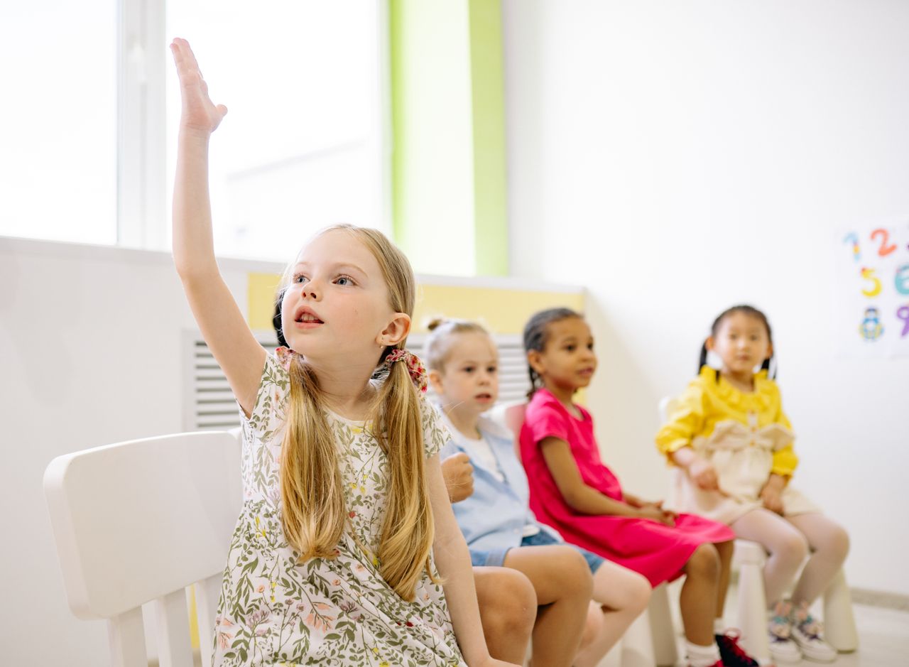 Kindergarten students in a classroom raising hands to participate in learning activities.